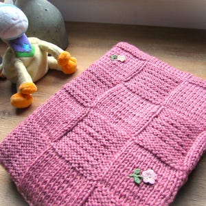 Könnte beinhalten: Eine handgestrickte, rosa Babydecke mit quadratischem Muster und kleinen floralen Verzierungen. Ein Plüsch-Giraffen-Spielzeug mit blauen und gelben Akzenten befindet sich im Hintergrund. Die Decke ist gefaltet und wirkt weich und gemütlich.