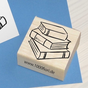 Könnte beinhalten: Ein quadratischer Holzstempel mit einer schwarzen Strichzeichnung eines Stapels aus drei Büchern und dem Text www.1000fiori.de, begleitet von einer weißen Klappkarte, die dieselbe schwarze Tuscheillustration der gestapelten Bücher zeigt.
