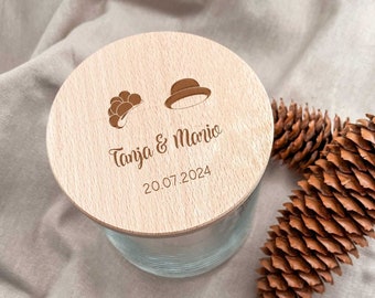 May include: A wooden lid with a light brown finish. The lid has a design of a hat and a flower with the text "Tanja & Mario 20.07.2024" engraved on it.