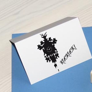 Op de afbeelding: Een witte gevouwen kaart en een houten stempel met een zwart koekoeksklokontwerp met het woord "Kuckuck!" en de website "www.1000fiori.de". De items liggen op een blauwe kaart, tegen een licht houten oppervlak.