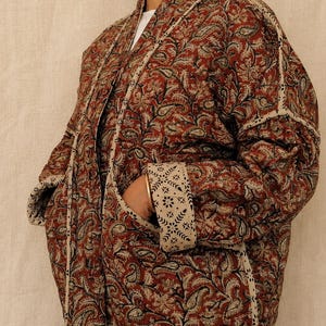 May include: A quilted jacket featuring a deep red and black paisley pattern on a cream background. The garment has an open front, long sleeves with folded cuffs, and contrasting cream-coloured trim with a small black geometric print.