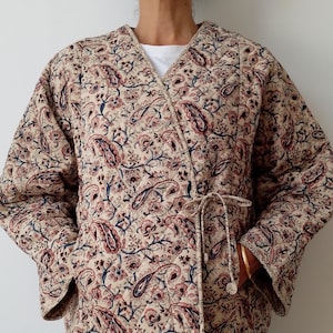 Puede incluir: Chaqueta acolchada beige con estampado de cachemir en tonos rojos, azules y marrones. La chaqueta cruzada tiene un lazo en la cintura y mangas largas. Prenda ligera con estampado floral.