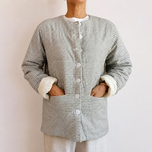 May include: A long-sleeved quilted jacket featuring a black and white grid pattern. The jacket has a round neckline, a front button closure with six white buttons, and two large patch pockets on the front. The sleeve cuffs are turned up to reveal a solid white interior lining.