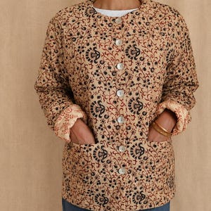 May include: A long-sleeved, quilted jacket in a beige hue featuring a repeating floral and vine pattern in black and dark red. The jacket has a round neckline, a front button closure with light-colored buttons, and two large patch pockets at the front.