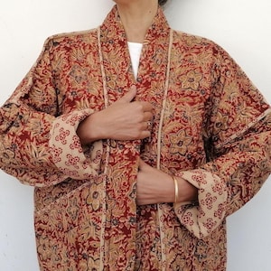 Op de afbeelding: Een kimono-achtige badjas met een rood en bruin bloemenpatroon. De badjas heeft een herhalend bloemenontwerp in tinten rood, bruin en beige. De mouwen en revers hebben een contrasterend beige bloemenpatroon. De badjas is open aan de voorkant en heeft een striksluiting.