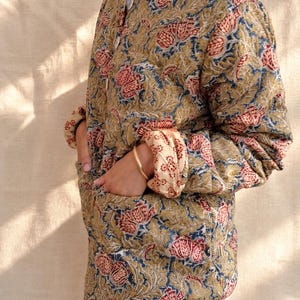 May include: A quilted jacket featuring a repeating floral pattern in shades of olive green, muted blue, and deep red. The jacket has a round neckline, a front button closure, long sleeves with folded cuffs, and two large patch pockets on the front.