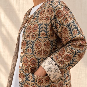 May include: A quilted jacket featuring a beige base with a repeating floral pattern in rust orange and dark blue. The jacket has a round neckline, long sleeves with folded cuffs revealing a white lining with black geometric dots, and white buttons.