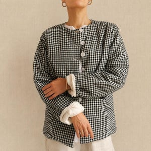 May include: A long-sleeved, quilted jacket featuring a black and white gingham check pattern. The jacket has a round neckline, a front button closure with white buttons, and white fabric cuffs visible at the wrists of the long sleeves.