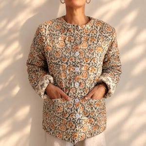 May include: A long-sleeved quilted jacket featuring a cream base with a repeating floral and vine pattern in shades of orange and dark blue. The jacket has a round neckline, a front button closure with five white buttons, and two front pockets.
