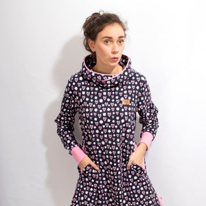 Puede incluir: Un vestido azul marino con un estampado de corazones rosas y blancos. El vestido tiene cuello de pico y mangas largas con ribetes rosas. El vestido tiene dos bolsillos y un cord&oacute;n en la parte inferior.