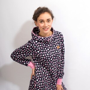Puede incluir: Un vestido azul marino con un estampado de corazones rosas y blancos. El vestido tiene cuello de pico y mangas largas con ribetes rosas. El vestido se muestra en una modelo con medias blancas.