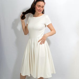Könnte beinhalten: Ein weißes Kleid mit vollem Rock und kurzen Ärmeln. Das Kleid hat ein schlichtes, elegantes Design und ist perfekt für verschiedene Anlässe. Das Model trägt ein rot-weiß gepunktetes Stirnband.