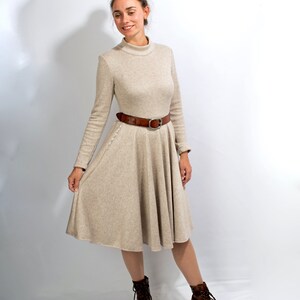 May include: A beige long-sleeved dress with a high neck and a brown belt. The dress has a flared skirt and the model is wearing brown ankle boots.