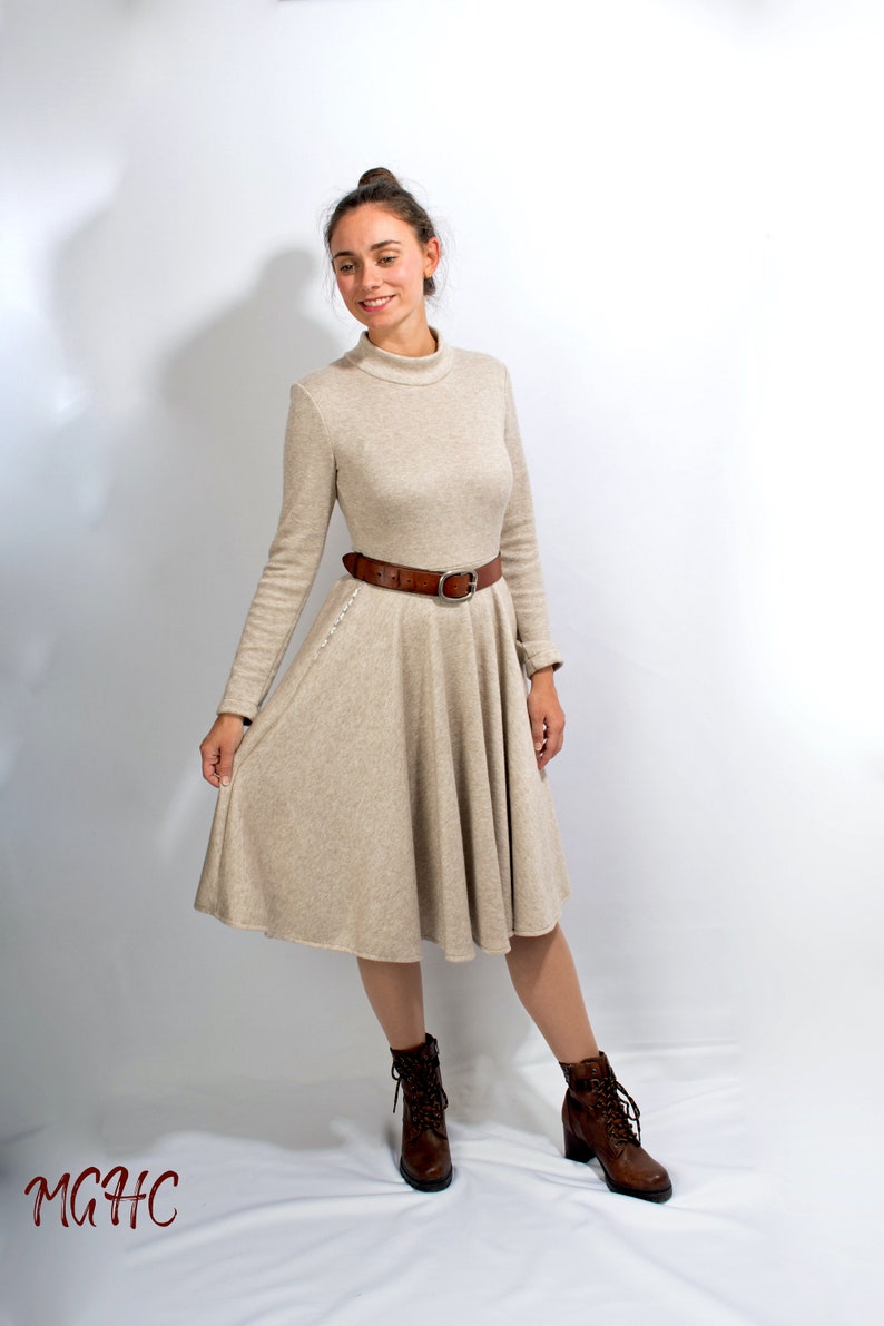 May include: A beige long-sleeved dress with a high neck and a brown belt. The dress has a flared skirt and the model is wearing brown ankle boots.