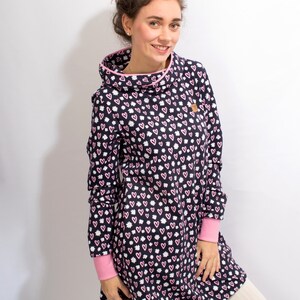 Puede incluir: Un vestido de manga larga con un estampado de corazones rosas y blancos sobre un fondo azul marino. El vestido tiene un cuello de pico y un ribete rosa alrededor del escote y los pu&ntilde;os. El vestido se muestra en una modelo que lleva leggings blancos.