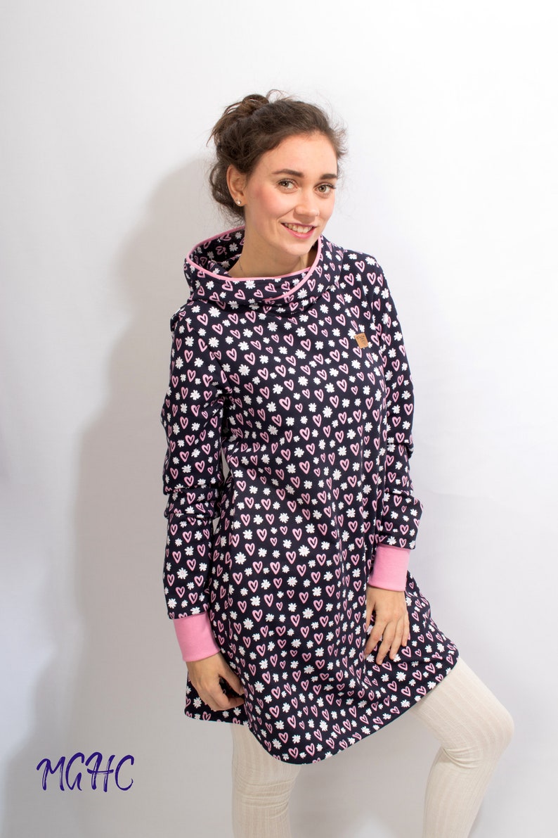 Puede incluir: Un vestido de manga larga con un estampado de corazones rosas y blancos sobre un fondo azul marino. El vestido tiene un cuello de pico y un ribete rosa alrededor del escote y los pu&ntilde;os. El vestido se muestra en una modelo que lleva leggings blancos.