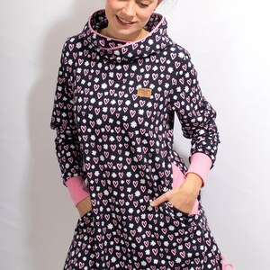 Puede incluir: Un vestido t&uacute;nica de manga larga de color azul marino con un estampado de corazones y flores rosas y blancas. El vestido tiene cuello de pico, mangas largas y dos bolsillos. El vestido tiene un cord&oacute;n de ajuste rosa en la parte inferior.