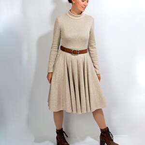 May include: A beige long-sleeved dress with a turtleneck and a brown belt. The dress has a flared skirt and the model is wearing brown lace-up boots.