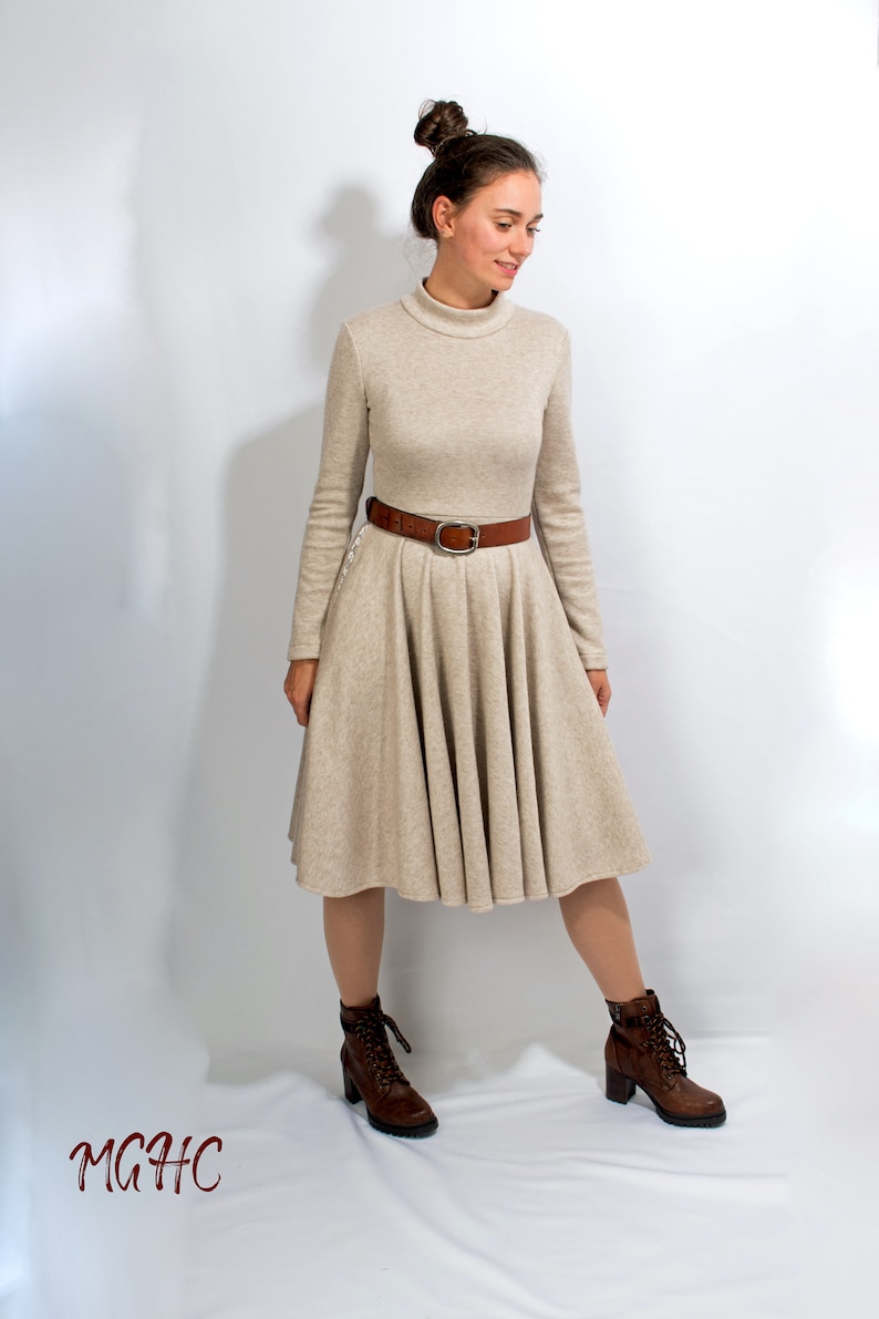 May include: A beige long-sleeved dress with a turtleneck and a brown belt. The dress has a flared skirt and the model is wearing brown lace-up boots.