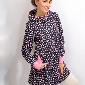 Puede incluir: Un vestido azul marino de manga larga con un estampado de corazones rosas y blancos. El vestido tiene un cuello de pico y un ribete rosa alrededor del escote. El vestido tiene un bolsillo en el lado izquierdo y un ribete rosa alrededor de los pu&ntilde;os.