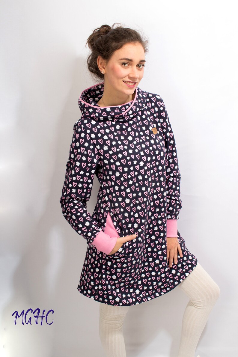 Puede incluir: Un vestido azul marino de manga larga con un estampado de corazones rosas y blancos. El vestido tiene un cuello de pico y un ribete rosa alrededor del escote. El vestido tiene un bolsillo en el lado izquierdo y un ribete rosa alrededor de los pu&ntilde;os.