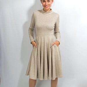 May include: A beige long-sleeved dress with a high neckline and a flared skirt. The dress has pockets on the sides and is made of a soft, comfortable fabric.
