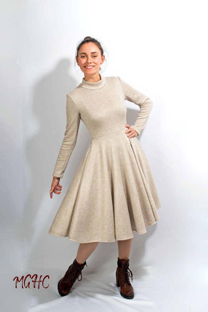 May include: A beige long-sleeved dress with a high neck and a flared skirt. The dress is made of a soft, knit fabric. The model is wearing brown ankle boots.