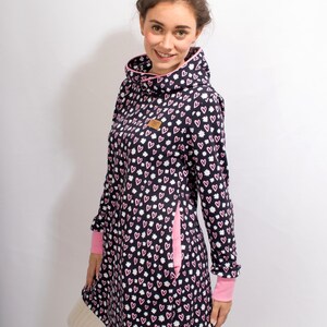Puede incluir: Un vestido de manga larga con un estampado de corazones rosas y blancos sobre un fondo azul marino. El vestido tiene un cuello alto y un ribete rosa. El vestido tiene un bolsillo lateral con un ribete rosa. El vestido tiene un cord&oacute;n en la parte inferior. La modelo lleva el vestido con medias blancas. El texto "MGHC" es visible en la esquina inferior derecha de la imagen.