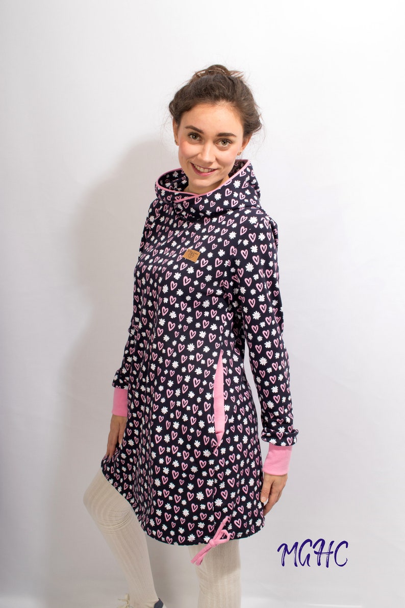 Puede incluir: Un vestido de manga larga con un estampado de corazones rosas y blancos sobre un fondo azul marino. El vestido tiene un cuello alto y un ribete rosa. El vestido tiene un bolsillo lateral con un ribete rosa. El vestido tiene un cord&oacute;n en la parte inferior. La modelo lleva el vestido con medias blancas. El texto "MGHC" es visible en la esquina inferior derecha de la imagen.