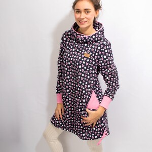 Puede incluir: Un vestido azul marino con ribete rosa y un estampado de corazones blancos y rosas. El vestido tiene cuello de pico y mangas largas. La modelo lleva medias blancas y zapatillas blancas.
