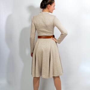 May include: A beige long-sleeved dress with a brown belt and a flared skirt. The dress is worn by a person with brown hair pulled back in a bun. The person is wearing brown boots.