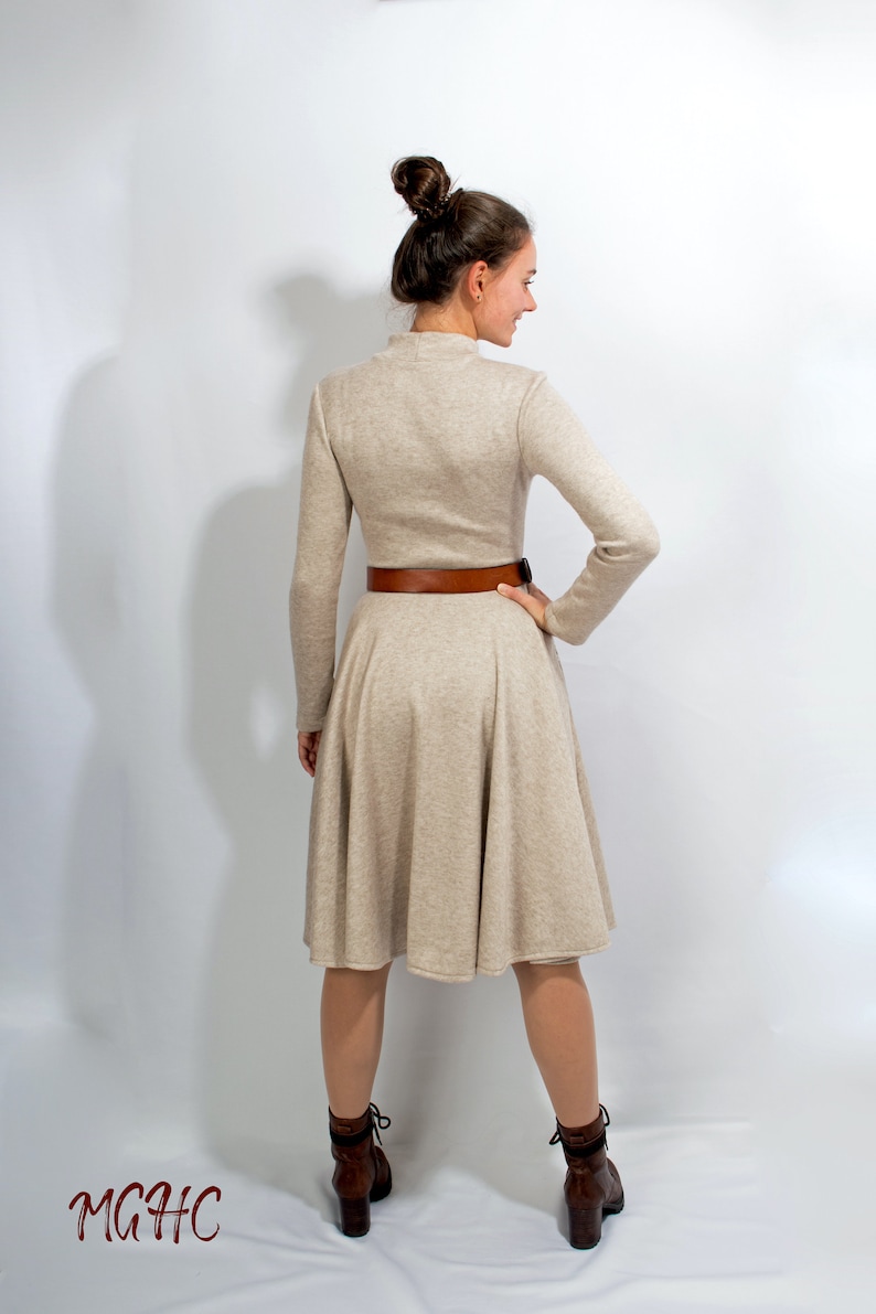 May include: A beige long-sleeved dress with a brown belt and a flared skirt. The dress is worn by a person with brown hair pulled back in a bun. The person is wearing brown boots.
