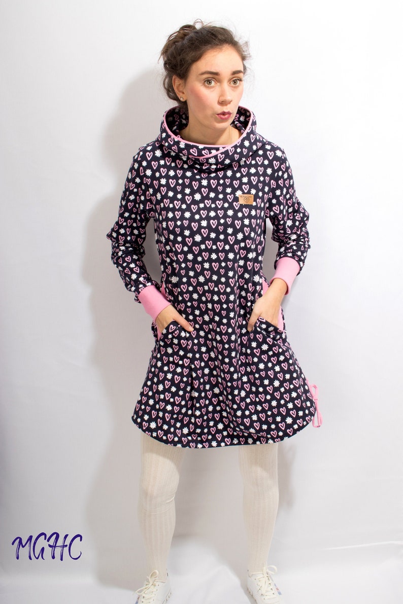 Puede incluir: Un vestido azul marino con un estampado de corazones rosas y blancos. El vestido tiene cuello de pico y mangas largas con ribetes rosas. El vestido tiene dos bolsillos y un cord&oacute;n en la parte inferior.