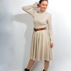 May include: A woman wearing a beige turtleneck dress with a brown belt and brown ankle boots. She is standing on a white background.