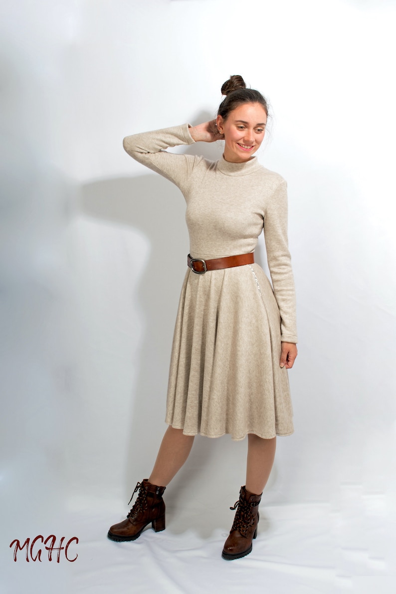 May include: A woman wearing a beige turtleneck dress with a brown belt and brown ankle boots. She is standing on a white background.