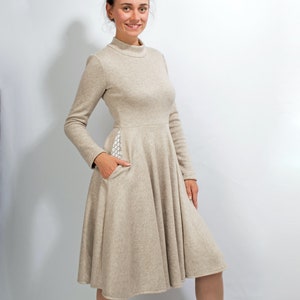 May include: A beige long-sleeved dress with a high neckline and a flared skirt. The dress has a small pocket on the side with a white patterned fabric. The model is wearing brown ankle boots.