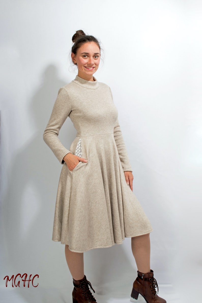 May include: A beige long-sleeved dress with a high neckline and a flared skirt. The dress has a small pocket on the side with a white patterned fabric. The model is wearing brown ankle boots.