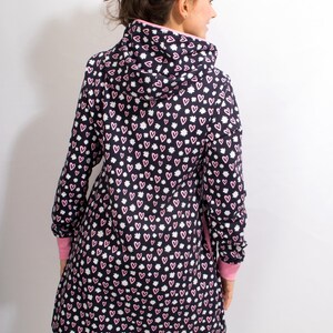 Puede incluir: Una chaqueta con capucha de manga larga con fondo negro y un patr&oacute;n de corazones rosas y flores blancas. La chaqueta tiene un ribete rosa alrededor de la capucha y los pu&ntilde;os.