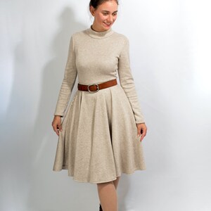 May include: A beige long-sleeved dress with a turtleneck and a brown belt. The dress has a flared skirt and the model is wearing brown ankle boots.