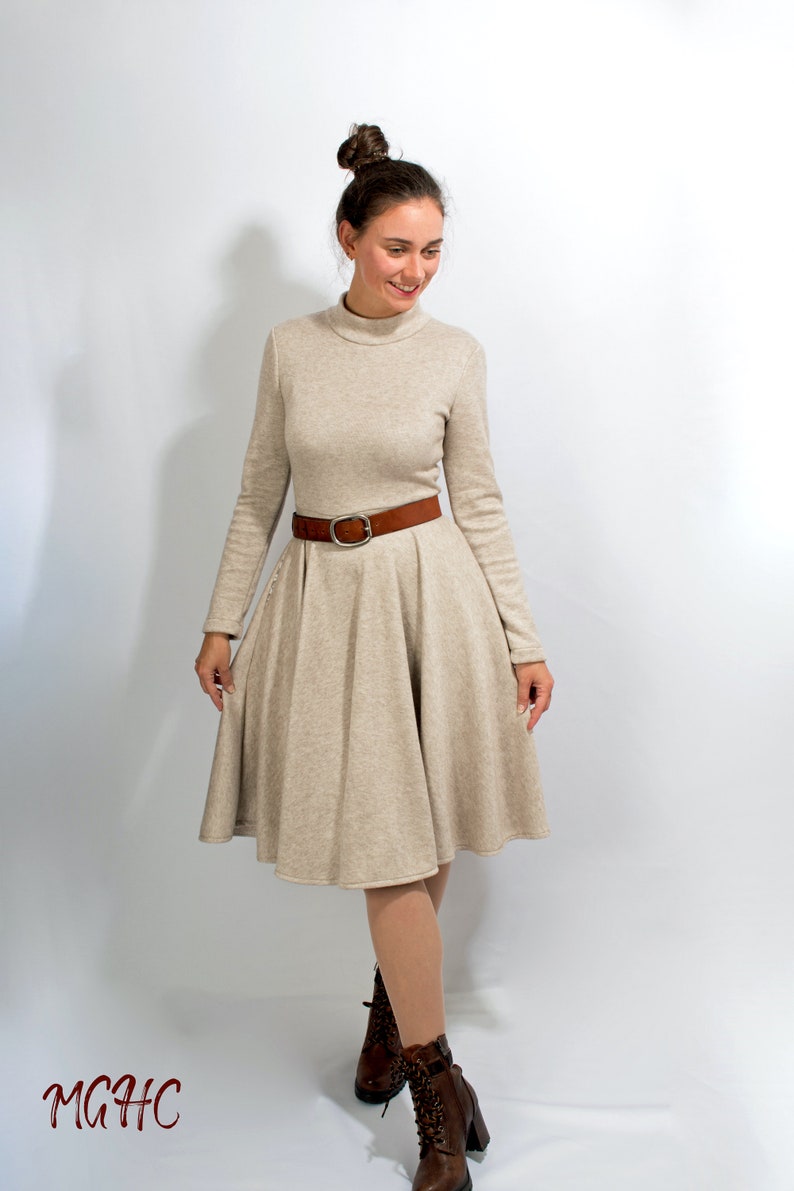May include: A beige long-sleeved dress with a turtleneck and a brown belt. The dress has a flared skirt and the model is wearing brown ankle boots.