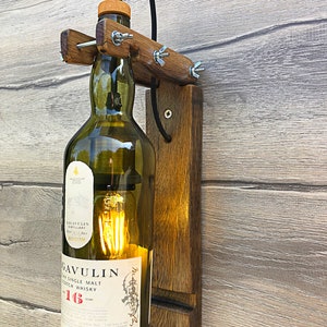 May include: A wall-mounted light fitting made from a repurposed whisky bottle. The bottle is clear glass with a brown label that reads "Lagavulin 16 Years Old Single Malt Scotch Whisky". The fitting is made of wood and has a rustic, industrial look.