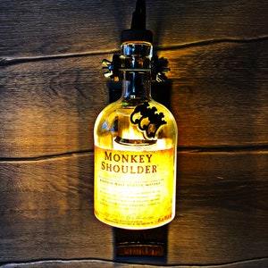 Puede incluir: Una botella de vidrio transparente con una fijación de metal negro y una luz en el interior. La botella está etiquetada con "Monkey Shoulder Blended Malt Scotch Whisky".