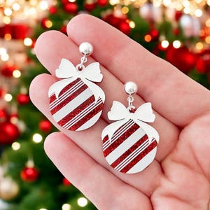 May include: A pair of Christmas-themed earrings. Each earring features a white bow and a round ornament design with red and white stripes. The earrings are held in a hand, with a blurred Christmas tree in the background.