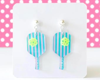 May include: A pair of pickleball paddle earrings. Each earring features a paddle shape with blue and white stripes, a yellow ball, and a silver stud. The earrings are displayed on a white background with a pink polka dot backdrop.