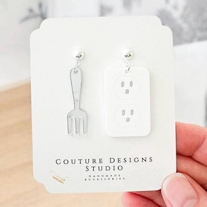 May include: A pair of quirky earrings, one shaped like a fork and the other like a white electrical outlet. The earrings are displayed on a white card with the text "COUTURE DESIGNS STUDIO HANDMADE ACCESSORIES".