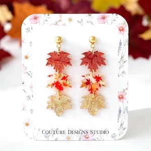 May include: Dangle earrings featuring three stacked maple leaf charms in red, white, and gold tones. The top leaf is textured red, the middle is white with red leaf patterns, and the bottom is textured gold. The earrings are displayed on a white card with floral accents and the text COUTURE DESIGNS STUDIO.
