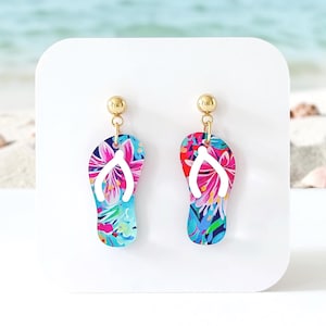 May include: A pair of dangle earrings featuring flat, flip-flop shaped charms. The charms display a vibrant, multi-colored floral pattern in shades of pink, blue, and green, with white straps. Each charm hangs from a small, polished gold-tone ball stud.