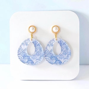 May include: A pair of teardrop-shaped earrings with a blue and white seashell pattern. The earrings have a gold-colored top with a small white pearl. The earrings are displayed on a white surface.