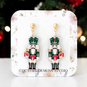 May include: A pair of gold-tone dangle earrings featuring nutcracker figures in green, red, and white enamel. The nutcrackers hold small red and white gift boxes. The earrings are displayed on a white card with floral accents and the text COUTURE DESIGNS STUDIO.