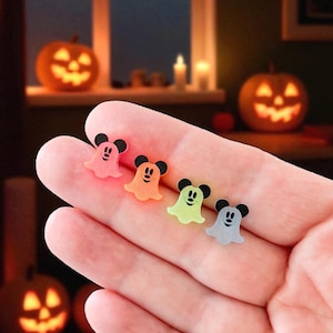 Puede incluir: Cuatro decoraciones de Halloween con forma de fantasma de colores con detalles negros de orejas de rat&oacute;n se sostienen en una mano. Los fantasmas son rosas, naranjas, verdes y grises. El fondo incluye calabazas iluminadas y una ventana.